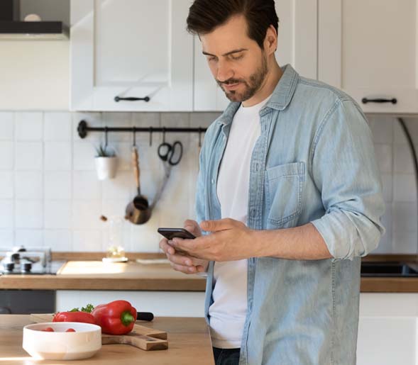 Man standing in a kitchen looking at his phone with a red bell pepper on a cutting block in front of him