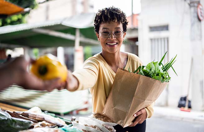 Woman purchasing a yellow bell pepper holding a brown bag of vegetables at a farmers market
